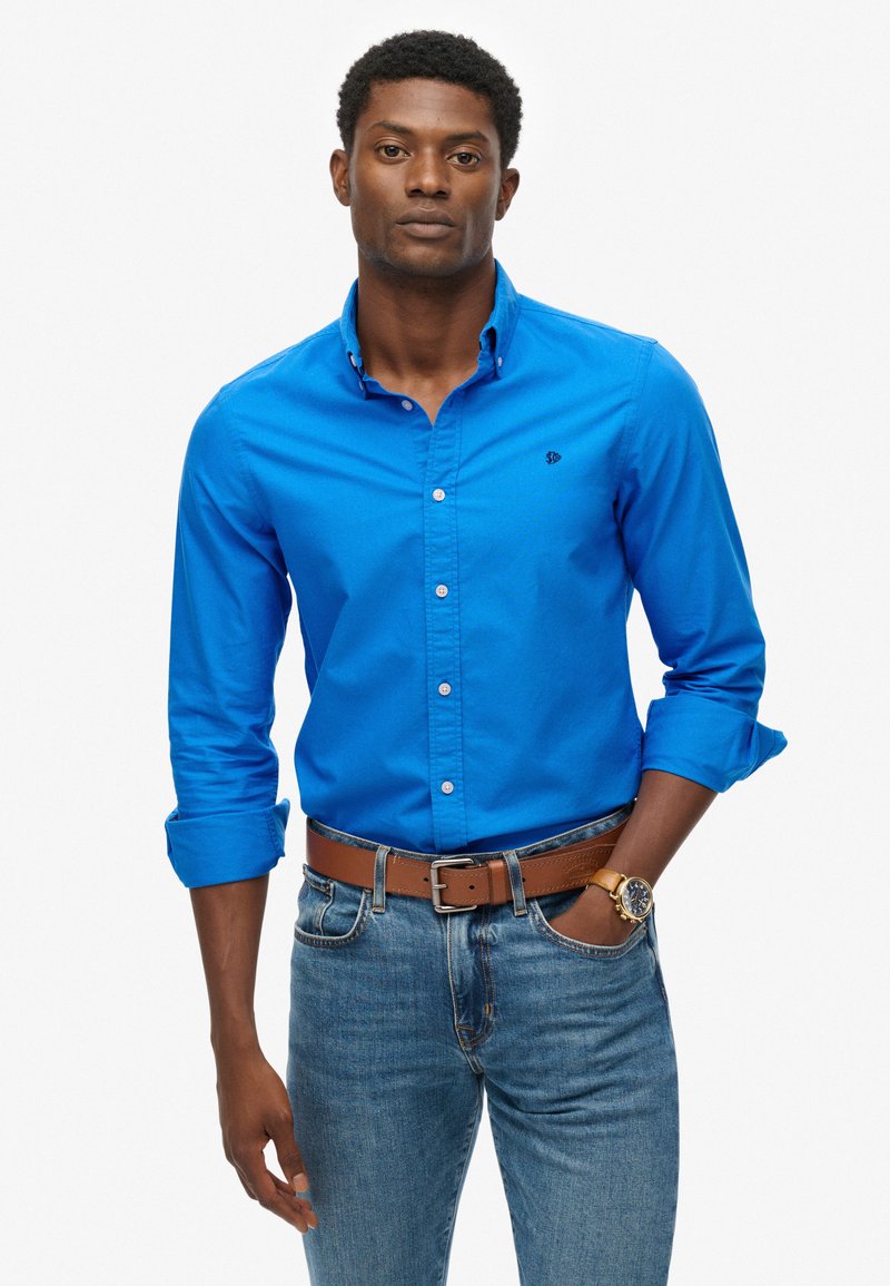 Blauw bloesje, van katoen, lange mouwen opgerold, klein logo op de borst, gecombineerd met een bruine riem en blauwe spijkerbroek.