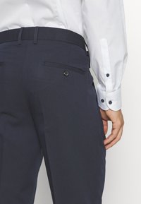 Dunkelblaue Hose mit glatter Textur, ausgestattet mit einer knöpfbaren Tasche und einer taillierten Passform, kombiniert mit einem weißen Hemd mit Knöpfen.