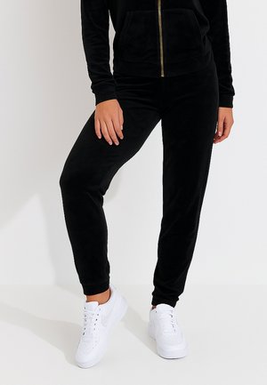 Schwarze Velours-Trainingshose mit schmal zulaufendem Bein, ausgestattet mit einem elastischen Bund und seitlichen Taschen, kombiniert mit weißen Sneakers.