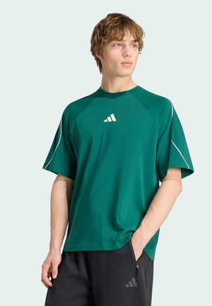 Ung mand iført grøn kortærmet sportsshirt og sorte træningsbukser med afslappede hænder, kiggende til siden på en ensfarvet baggrund.