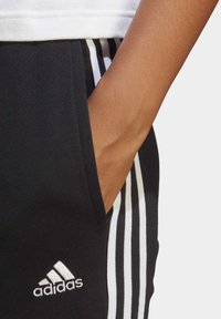 Svarta träningsbyxor med vit tre-stripes-design, med sidofickor och en liten vit Adidas-logotyp på nedre vänstra benet.