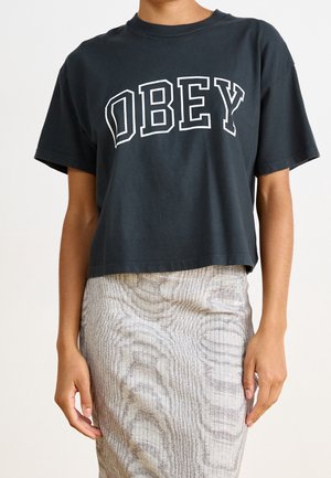 Person iført en sort kort T-shirt med teksten "OBEY" og et beige nederdel med høj talje med et subtilt abstrakt mønster.
