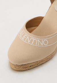 Bež platnena espadrila z zaobljenim nosom, ki ima bel vezen logotip in pleteno juten podplat. Gladka tekstura, minimalni oblikovni detajli.