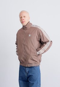 Brun zip-up jakke med høy krage, med hvite striper på ermene og et brodert logo på brystet. Dongeribukser nedenfor.
