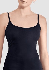 Mujer vistiendo una camiseta negra ajustada con tirantes finos y aretes en forma de corazón frente a un fondo claro y sencillo.