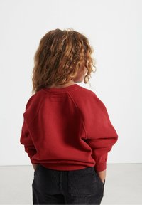 Karmyno raudonas džemperis su laisvu siluetu ir megztais rankogaliais, derinamas su tamsiais šamono kelnais. Atgalinė pusė, rodanti raglan rankoves ir tekstūruotą audinį.