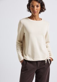 Femme avec les cheveux courts et bouclés, portant un haut beige à manches longues avec un nœud sur le côté et un pantalon marron foncé, les mains dans les poches, arrière-plan neutre.