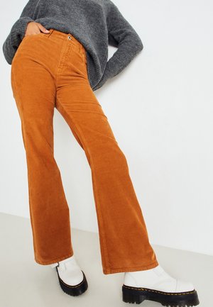 Osoba ubrana w spodnie sztruksowe w kolorze spalanej pomarańczy, szary sweter i białe buty na platformie, stojąca przy białej ścianie.