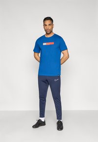 Μπλε κοντομάνικο μπλουζάκι με κόκκινο και κίτρινο λογότυπο «BARCA»· συνδυασμένο με σκούρα μπλε παντελόνια jogger που φέρουν λευκό σήμα Nike· μαύρα αθλητικά παπούτσια.