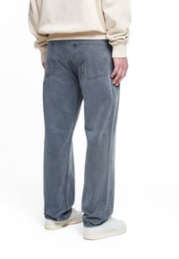 Asmuo, vilkintis laisvus pilkus džinsus ir baltus sportinius batelius, apsirengęs kreminės spalvos megztiniu, stovi pasisukęs atgal prieš baltą foną.