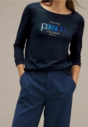 Mujer con camisa de manga larga azul marino con el texto "inspiring passion time ahead" y pantalones azul marino, con las manos en los bolsillos, fondo neutro.