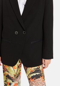 Black blazer with double buttons, side pockets, and satin trims, paired with colorful patterned pants featuring animal and floral designs.