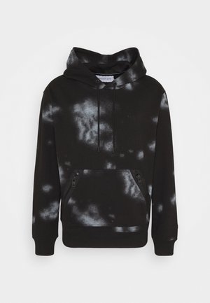 Osoba ubrana w czarno-białą bluzę z kapturem w stylu tie-dye, białe spodnie dresowe i czarne trampki z turkusowymi detalami, stoi z rękami w kieszeniach.