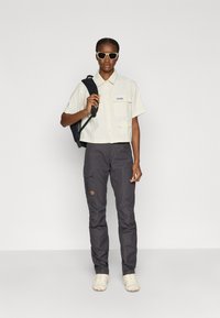 Kortärmad krämfärgad skjorta med knappar och ficka, kombinerad med mörkgrå cargobyxor med lapp och beige skor. Ryggsäck på axeln.