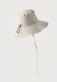 Φαρδύ καπέλο ήλιου σε ανοιχτό μπεζ χρώμα με ρυθμιζόμενο μαύρο κορδόνι και λουράκι για το πηγούνι, απεικονίζεται σε απλό λευκό φόντο.