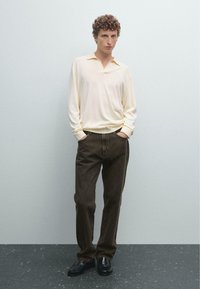 Ljust beige långärmad skjorta med v-ringning, mörkbruna byxor och svarta loafers, stående mot en ljusgrå bakgrund.