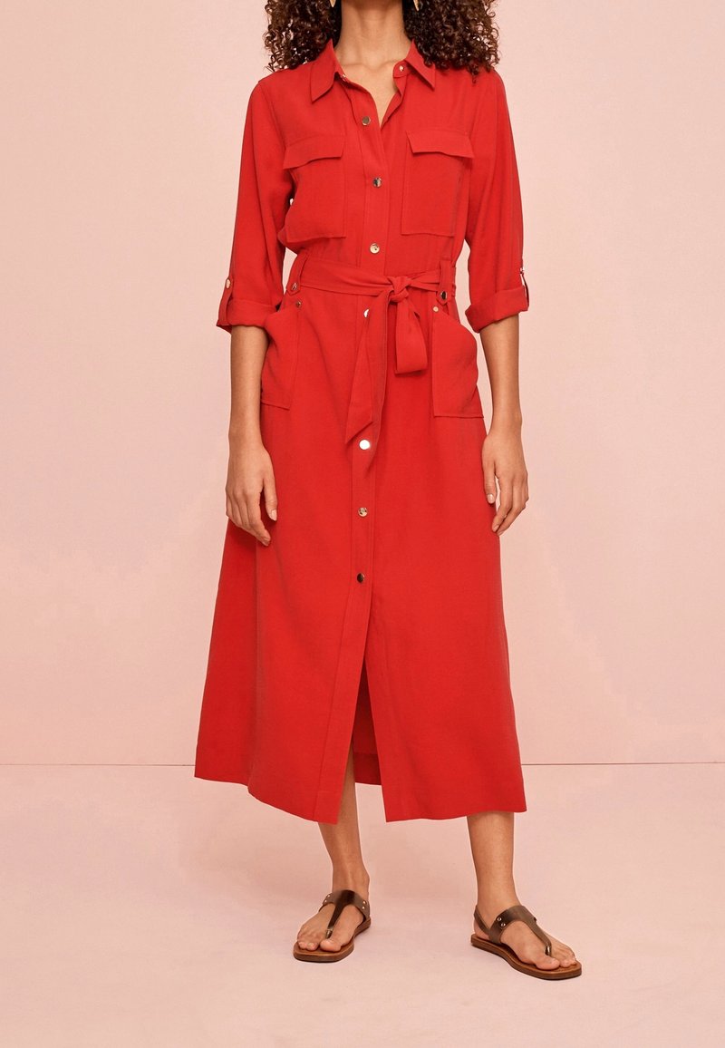 Femme portant une longue robe rouge à boutons avec des manches retroussées et une taille cintrée, associée à des sandales plates marron foncé sur un fond uni.