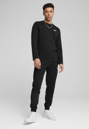 Schwarzes Langarmshirt mit Logo auf der Brust, kombiniert mit passenden schwarzen Jogginghosen und weißen Turnschuhen. Das Material wirkt glatt; taillierte Passform.