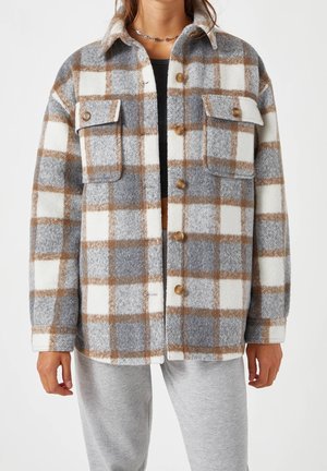 Kobieta ubrana w szarą, białą i brązową kraciastą kurtkę zapinaną na guziki z kieszeniami na piersi, na czarnej bluzce i szarych spodniach dresowych.