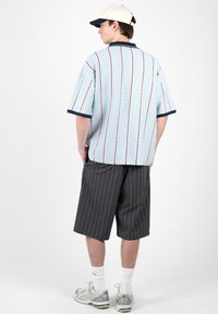 Homme de dos portant une chemise bleu clair à manches courtes avec des rayures rouges, un short foncé à fines rayures, des chaussettes blanches, des baskets et une casquette de baseball beige.