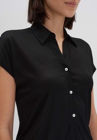 Camisa negra de manga corta con cuello, con un cierre de botones en la parte delantera y una textura suave, ligeramente brillante.