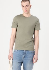 Ελαιοπράσινο βαμβακερό T-shirt με κοντά μανίκια, στρογγυλή λαιμόκοψη, λεία υφή. Συνδυασμένο με ανοιχτό μπλε τζιν και κρατώντας μια μπεζ τσάντα.