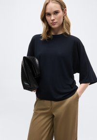 Γυναίκα με ξανθά μαλλιά μέχρι τους ώμους, φορώντας ένα χαλαρό μαύρο τοπ και μπεζ παντελόνι, κρατώντας μια μεγάλη μαύρη τσάντα φάκελο με υφή.