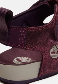 Gros plan sur le talon d'une sandale bordeaux avec une sangle ajustable, un matériau texturé et un logo d'arbre blanc sur la semelle intérieure et le côté extérieur.