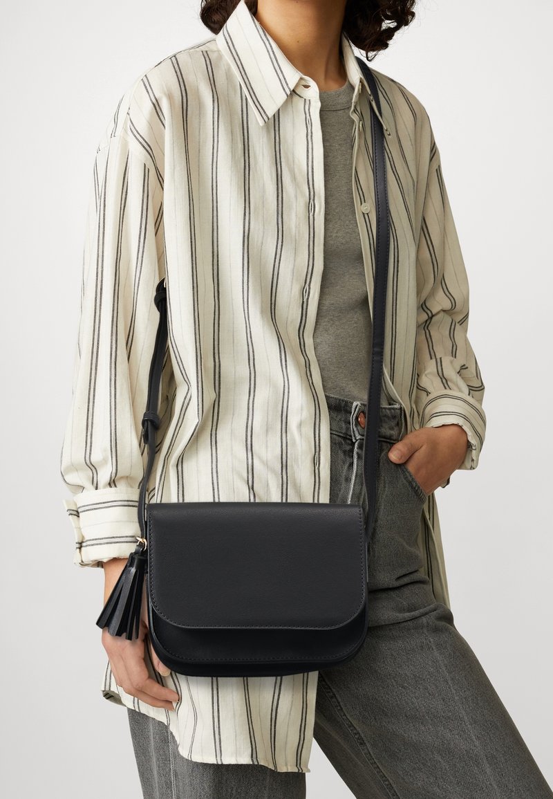 Borsa a tracolla in pelle nera con chiusura a pattina e dettaglio a nappina. La borsa ha una forma rettangolare slim e una tracolla regolabile.