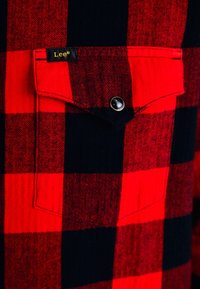 Rot- und schwarzgestreiftes Flanellhemd mit einer einzelnen Brusttasche, das ein schwarzes Etikett mit goldener Schrift und einen Druckknopfverschluss aufweist.