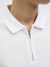 Witte poloshirt met een gestructureerde stof, voorzien van een ritssluiting aan de voorkant en een vlakke kraag. Het ontwerp is minimalistisch en eigentijds.