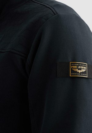 Črna majica z dolgimi rokavi, ki ima prešite detajle na rami in črno nalepko z zlatim logotipom "PME Legend American Classic" na rokavu.