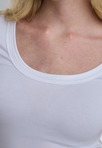 Personne blonde portant un t-shirt blanc à encolure dégagée, montrant le haut de la poitrine et la zone de la clavicule sur un fond neutre.