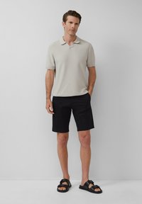 Beige strukturiertes Poloshirt mit kurzen Ärmeln, kombiniert mit schwarzen Shorts und schwarzen Sandalen mit einer Schnalle.
