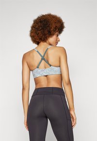 Top desportivo cinza e azul claro com um padrão texturizado, apresentando alças cruzadas e fechos ajustáveis. Combinado com leggins pretas.
