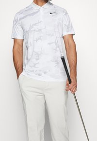 Muž v bílo-šedém maskáčovém polo tričku s krátkým rukávem a světle béžových kalhotách, držící golfovou hůl v jedné ruce.