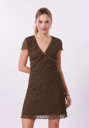 Junge Frau in einem braunen Spitzekleid mit kurzen Ärmeln, V-Ausschnitt und Schleifendetail, vor einem schlichten hellen Hintergrund stehend.