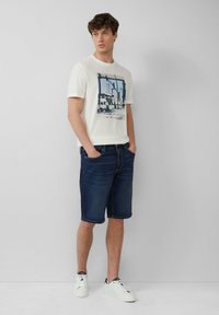 T-shirt en coton à col rond couleur écru avec un imprimé graphique, associé à un short en denim noir et des baskets blanches. Tenue décontractée avec poches.