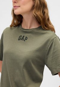 Olívazöld pamut póló kerek nyakkivágással és rövid ujjakkal, a mellkas felett kontrasztos fekete "GAP" felirattal.