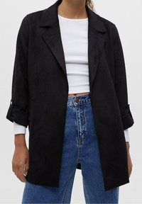 Black suede-like long coat with a collar, rolled sleeves, and open front; paired with a white top and high-waisted blue jeans.