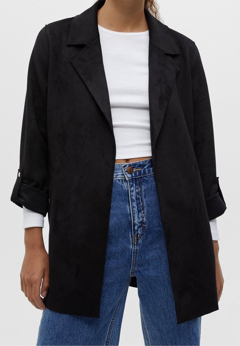 Black suede-like long coat with a collar, rolled sleeves, and open front; paired with a white top and high-waisted blue jeans.