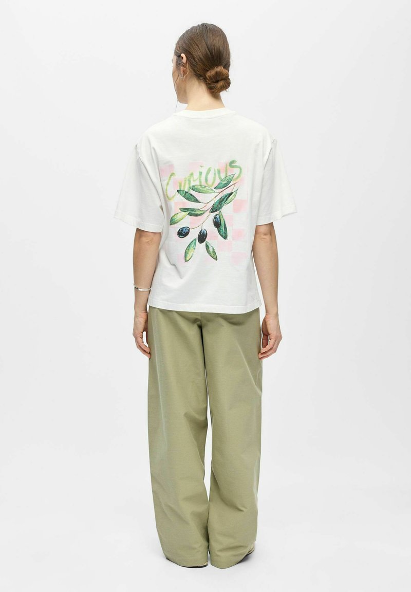 Femme aux cheveux en chignon portant un t-shirt blanc avec une branche d'olivier verte et le texte « Curious » dans le dos, associé à un pantalon ample vert olive.