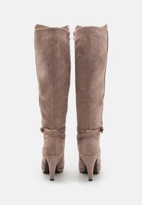 Botas de ante en color topo hasta la rodilla, con un tacón delgado. Cuentan con una cremallera lateral y una correa decorativa con herrajes dorados en el tobillo.