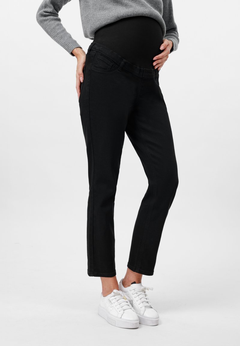Jeans de maternidad negros con una cinturilla elástica alta, forma ajustada y costuras en contraste. Llevados con un suéter gris claro y zapatos blancos.