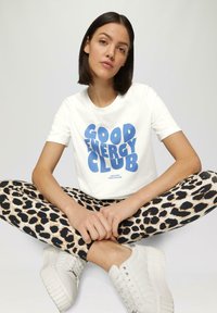 Witte t-shirt met blauwe tekst "GOOD ENERGY CLUB", gecombineerd met zwarte legging met een luipaardprint. Witte sneakers met een textuurdesign.