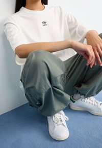 Bílá bavlněná tričko s černým logem Adidas, volné zelené kalhoty a bílé tenisky se zelenými detaily, sedící na modrém koberci.