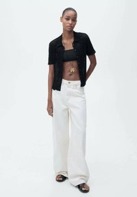 Cardigan noir à manches courtes, en tricot texturé, avec fermeture par boutons, superposé à un crop top noir. Associé à un jean blanc à jambes larges et des sandales noires.