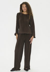 Donna con capelli ricci che indossa un top a maniche lunghe a coste marrone scuro e pantaloni a gamba larga abbinati, in piedi a piedi nudi su uno sfondo bianco.