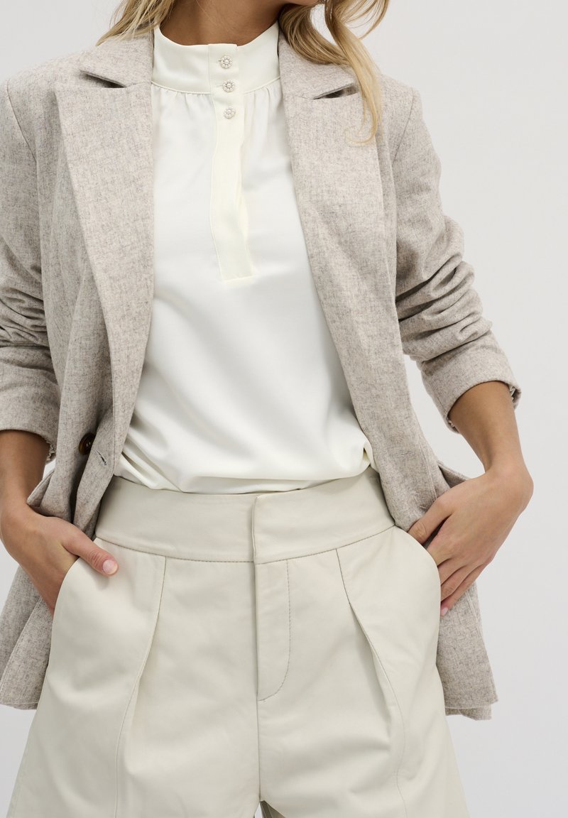 Femme portant un blazer beige cintré, un chemisier blanc à boutons décoratifs et un pantalon taille haute crème, mains dans les poches, fond neutre.