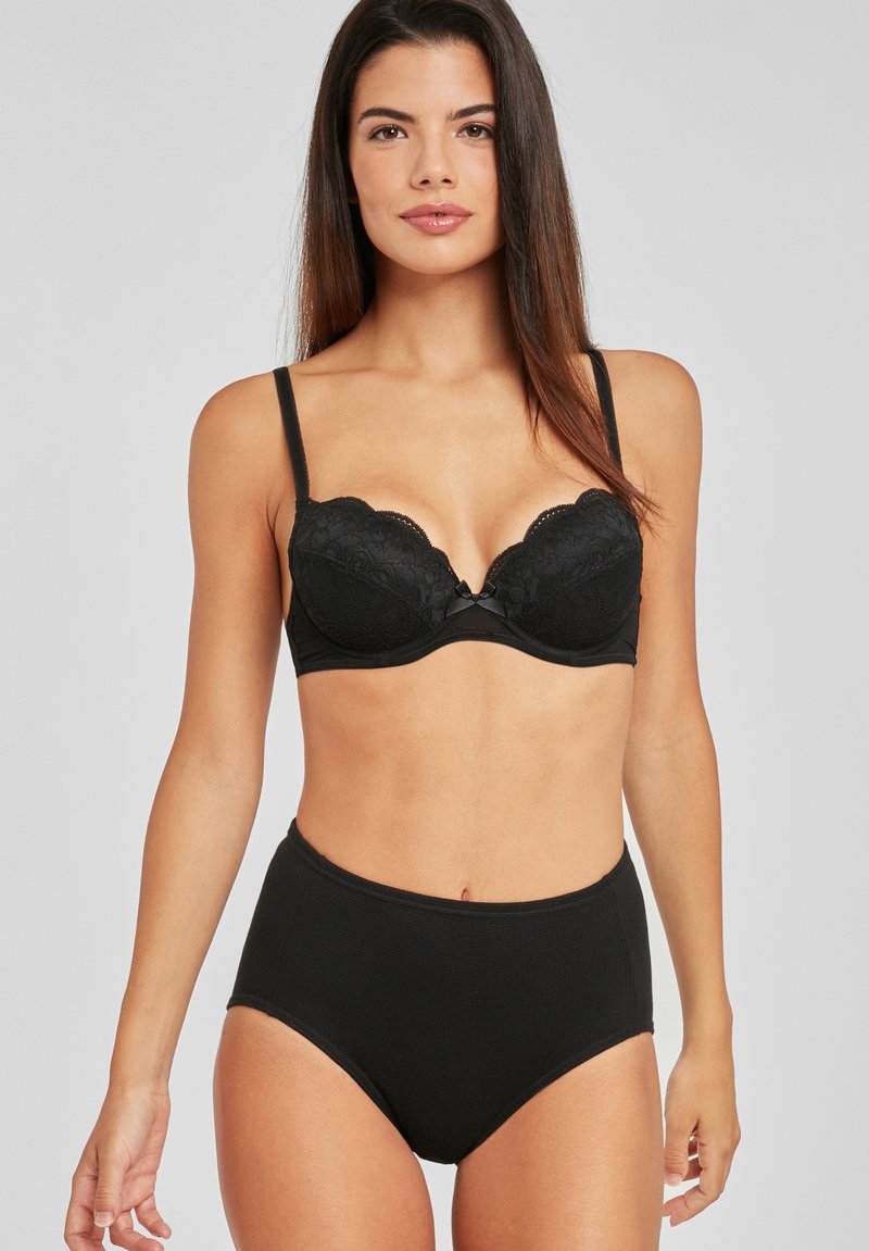 Melnas mežģīņu krūšturis ar polsterētām krūšu kausām, viļņotām malām un regulējamām siksnām, apvienots ar melnām kokvilnas biksēm ar augstu jostasvietu.